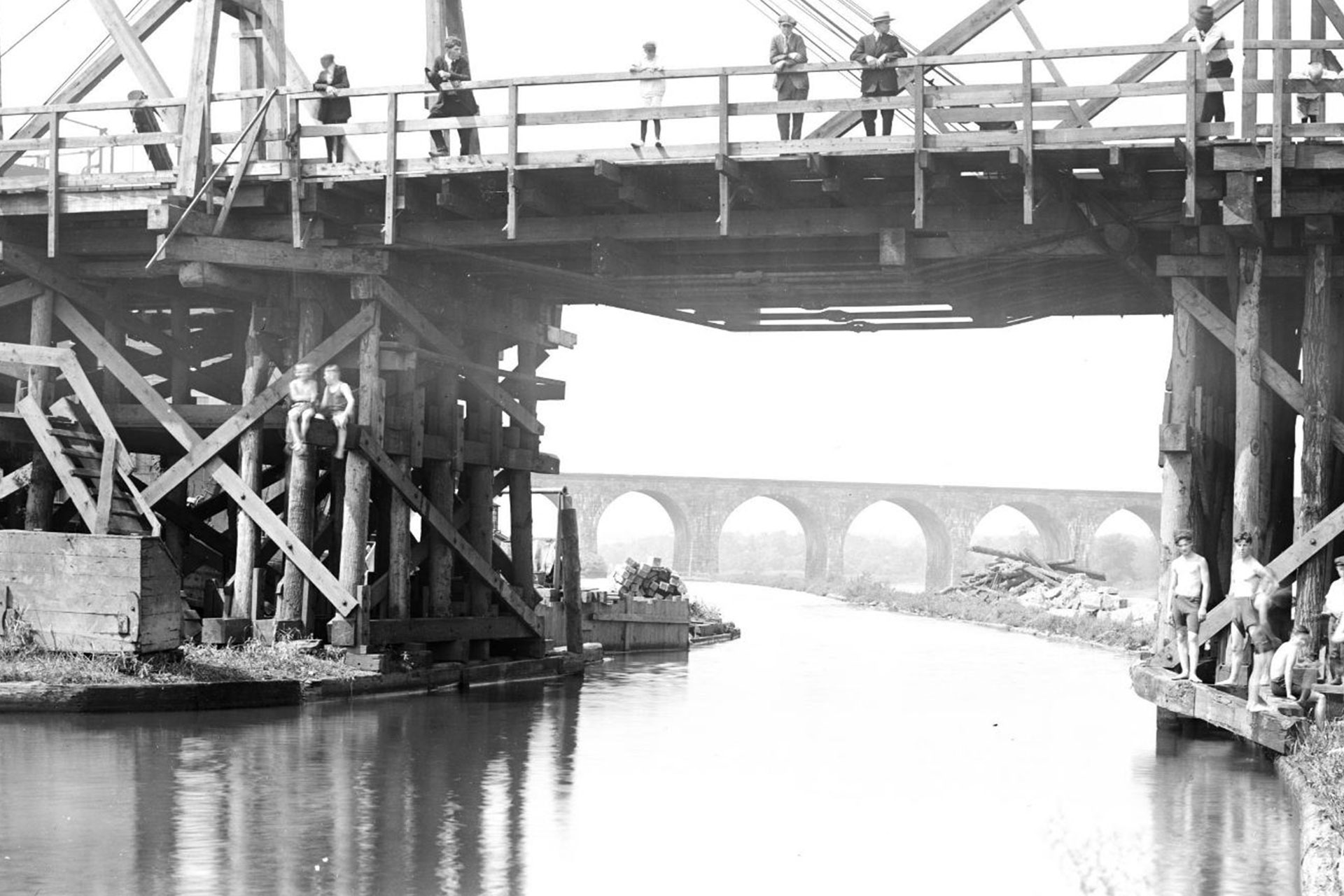 On a hot summer’s day, youngsters swim from a bridge in New Brunswick while onlookers watch from above. The arched bridge in the background carried the Pennsylviania RR over the D&R Canal and Raritan River.