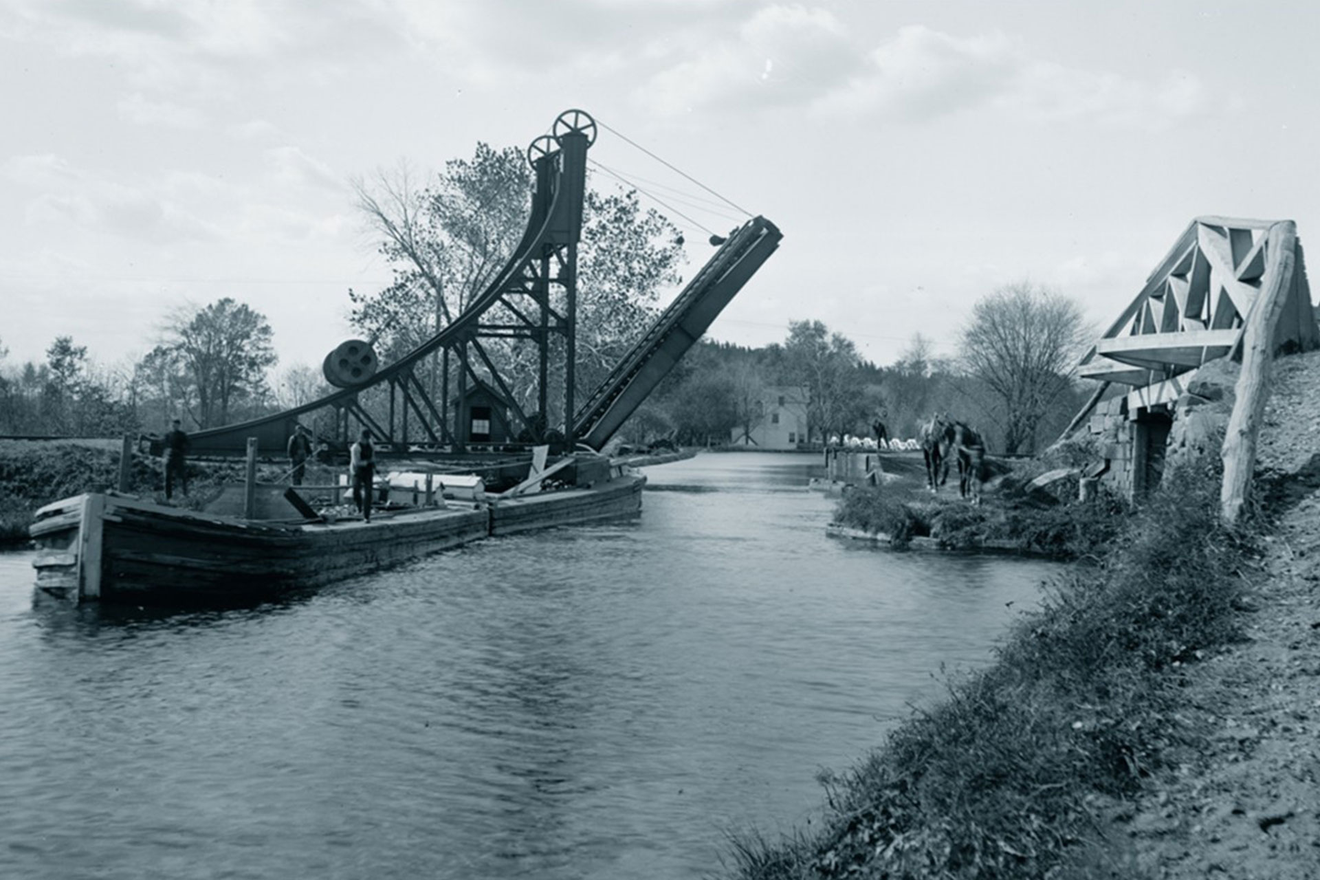 A loaded boat heading east has just passed under the drawbridge of the NY and Greenwood Lake RR. The bridge over the entrance to the Pompton Feeder is on the right. In the distance is the Pompton River aqueduct.