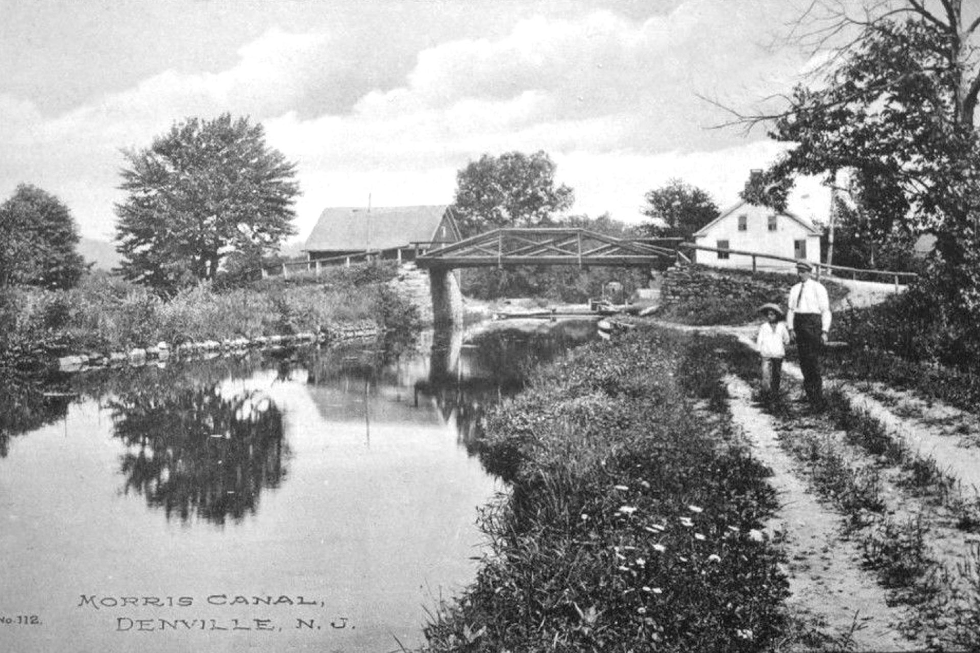 A man and child stand on the towpath, with Peer’s canal store and Diamond Spring Road bridge in the background in Denville.